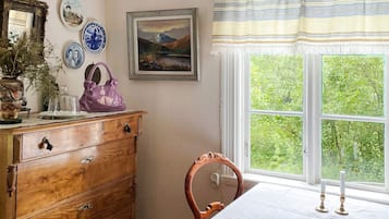 A room with a wooden dresser, a chair, a table with a white tablecloth, a window with a view of greenery, and decorative plates and a framed picture on the wall.
