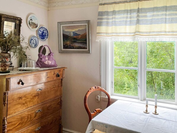 A room with a wooden dresser, a chair, a table with a white tablecloth, a window with a view of greenery, and decorative plates and a framed picture on the wall.