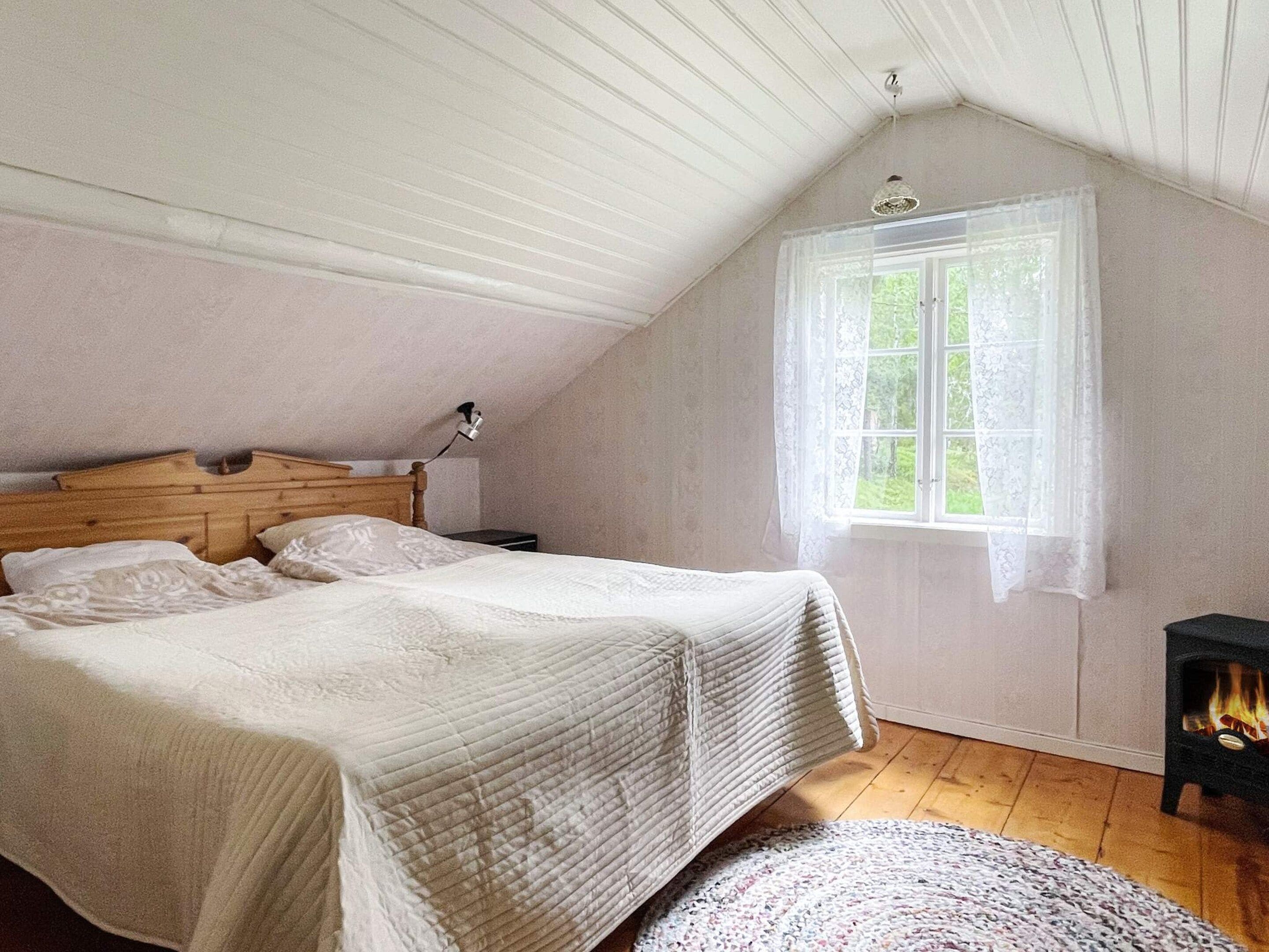 Un dormitorio con techo inclinado, una cama con cabecera de madera, una mesita de noche, un ventanal con cortinas de encaje y una chimenea.