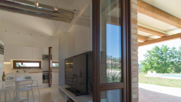 Una cuina moderna amb armaris blancs, un televisor gran i una zona de menjador amb taula i cadires de vidre.