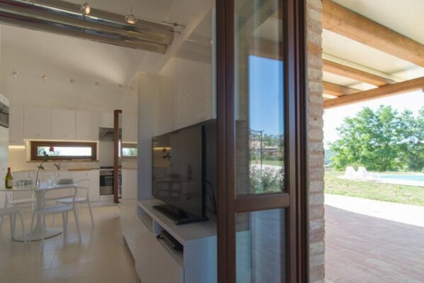 Una cocina moderna con armarios blancos, un televisor grande y un comedor con mesa y sillas de vidrio.