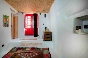 Quarto com teto de madeira, porta vermelha, um pequeno banquinho e tapete estampado.
