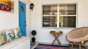 Een kamer met een blauwe deur, een rieten stoel, een klein tafeltje en een blauw gestreept vloerkleed.