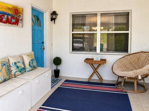 Ein Zimmer mit blauer Tür, einem Korbstuhl, einem kleinen Tisch und einem blau gestreiften Teppich.