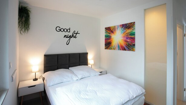 Ein Schlafzimmer mit einem Bett, zwei Nachttischen, einer an der Wand befestigten Pflanze, einem Schild mit der Aufschrift 'Good night' und einem farbenfrohen abstrakten Gemälde.