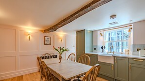 Un comedor con mesa y sillas de madera, un jarrón con flores, y una cocina con armarios verdes y una ventana.