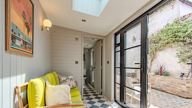Una habitación con piso a cuadros, un sofá verde con cojines, un cuadro en la pared y vistas a un espacio exterior con pared de ladrillo y vegetación a través de una puerta de vidrio.