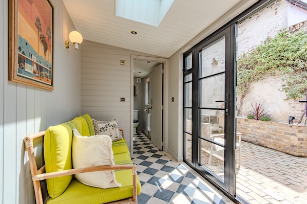 Ein Zimmer mit Schachbrettmuster-Boden, einem grünen Sofa mit Kissen, einem Bild an der Wand und Blick durch eine Glastür auf einen Außenbereich mit Ziegelwand und Grünpflanzen.