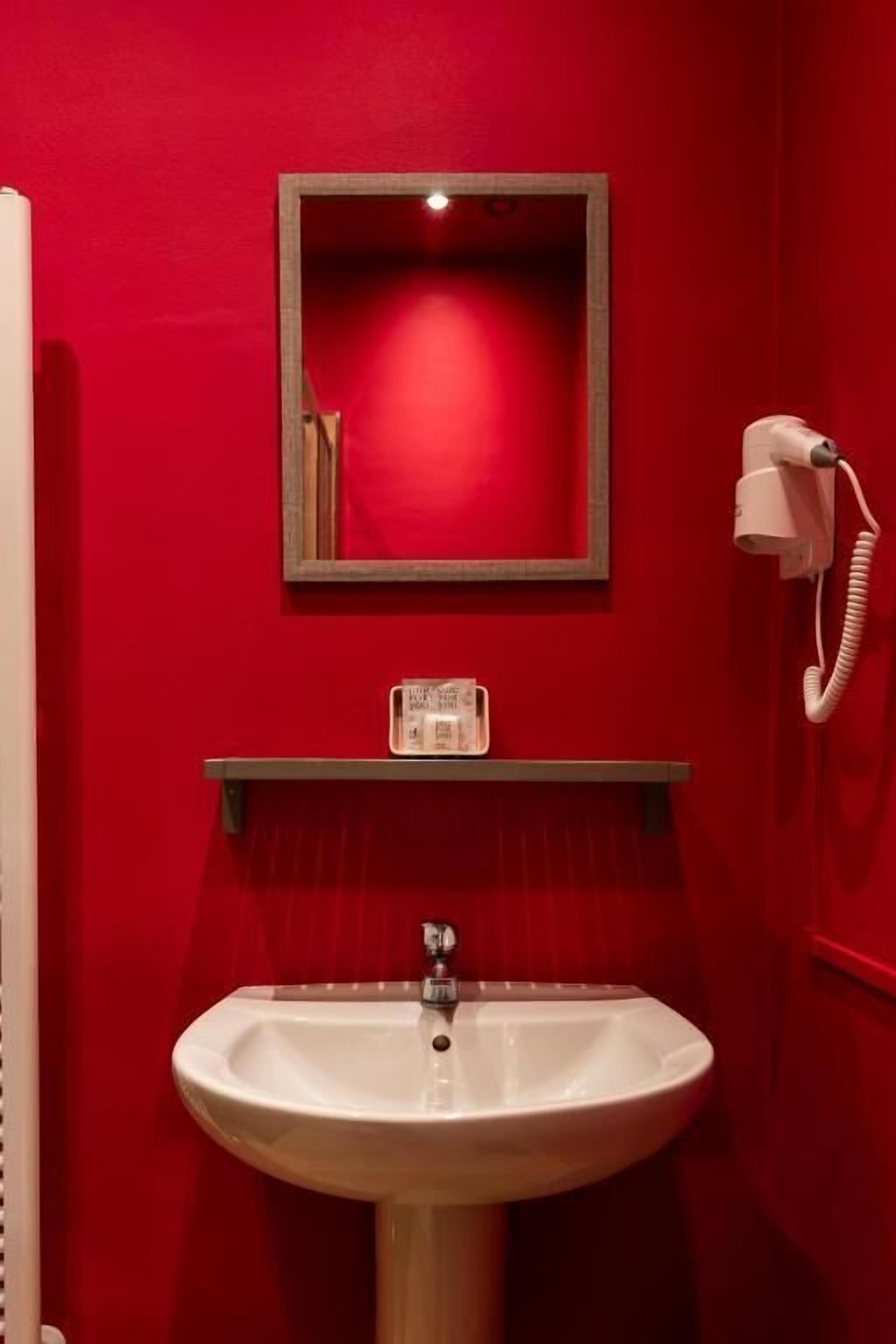 Ein Badezimmer mit roter Wand, Waschbecken in Weiß, einem Spiegel und einem Telefon.