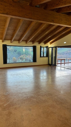 Una habitación amplia con techo alto, ventanas grandes con cortinas, piso de madera y vista a un paisaje boscoso.
