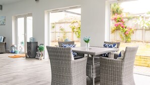 Un patio con mobiliario de mimbre, una mesa y vistas a un jardín trasero con valla de madera y plantas en flor.