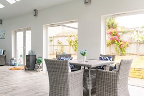 Un patio con mobiliario de mimbre, una mesa y vistas a un jardín trasero con valla de madera y plantas en flor.
