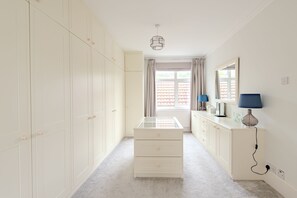 A room with a walk-in closet, a glass-top dresser, a lamp, and a window with curtains.