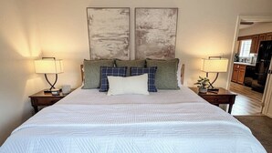 Quarto com cama grande, duas mesas de cabeceira, um criado-mudo, luminária na parede e porta de acesso a um banheiro.