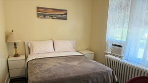 Un dormitorio con cama, mesita de noche, lámpara, radiador y ventana con cortinas.