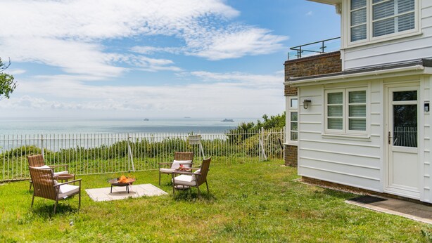 A white building with a balcony, a grassy area with outdoor furniture, and a view of the sea.