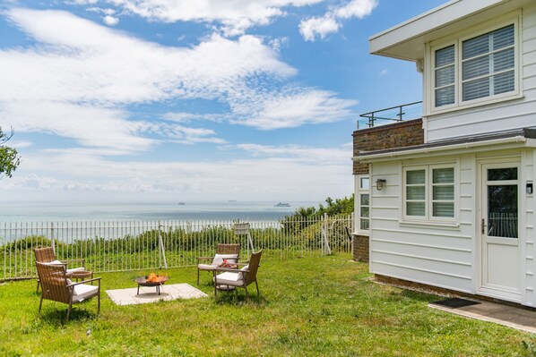 A white building with a balcony, a grassy area with outdoor furniture, and a view of the sea.
