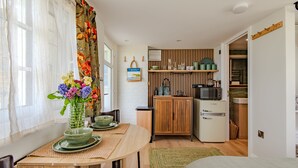 A compact kitchen with a microwave, refrigerator, and wooden shelves. A dining area with a table and chairs is visible, featuring a vase with flowers.