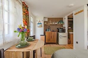 A compact kitchen with a microwave, refrigerator, and wooden shelves. A dining area with a table and chairs is visible, featuring a vase with flowers.
