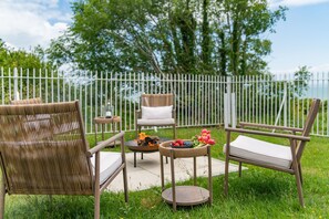 An outdoor seating area with wicker chairs and a table, surrounded by a white fence and greenery.