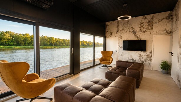 A modern living room with a large window overlooking a body of water, a flat-screen TV mounted on the wall, and a seating area with leather sofas and chairs.