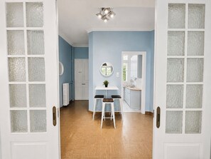 A hallway with a wooden floor, a white door with frosted glass panels, a white wall with a round mirror, and a small table with two chairs. - The Acropolis Neighbourhood Experience Apt by ΜΡS (Athina)