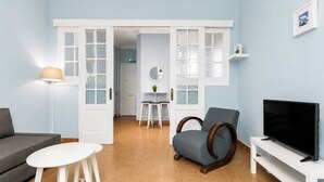 Ein Wohnzimmer mit grauem Sofa, einem weißen Couchtisch, einem blauen Sessel, einem Fernseher auf einem weißen Schrank und einer Tür zu einem anderen Raum.