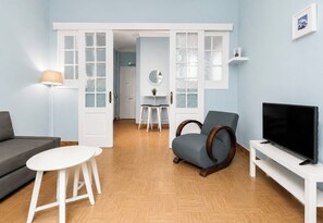 A living room with a grey sofa, a white coffee table, a blue armchair, a TV on a white stand, and a door leading to another room. - The Acropolis Neighbourhood Experience Apt by ΜΡS (Athina)