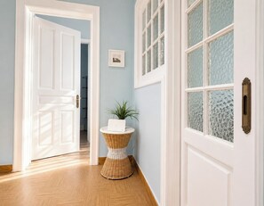 A hallway with a white door, a wicker side table with a plant, and a window with textured glass. - The Acropolis Neighbourhood Experience Apt by ΜΡS (Athina)