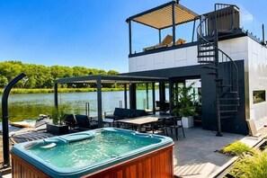 A modern outdoor hot tub area with a pool, lounge chairs, and a covered seating area overlooking a lake.