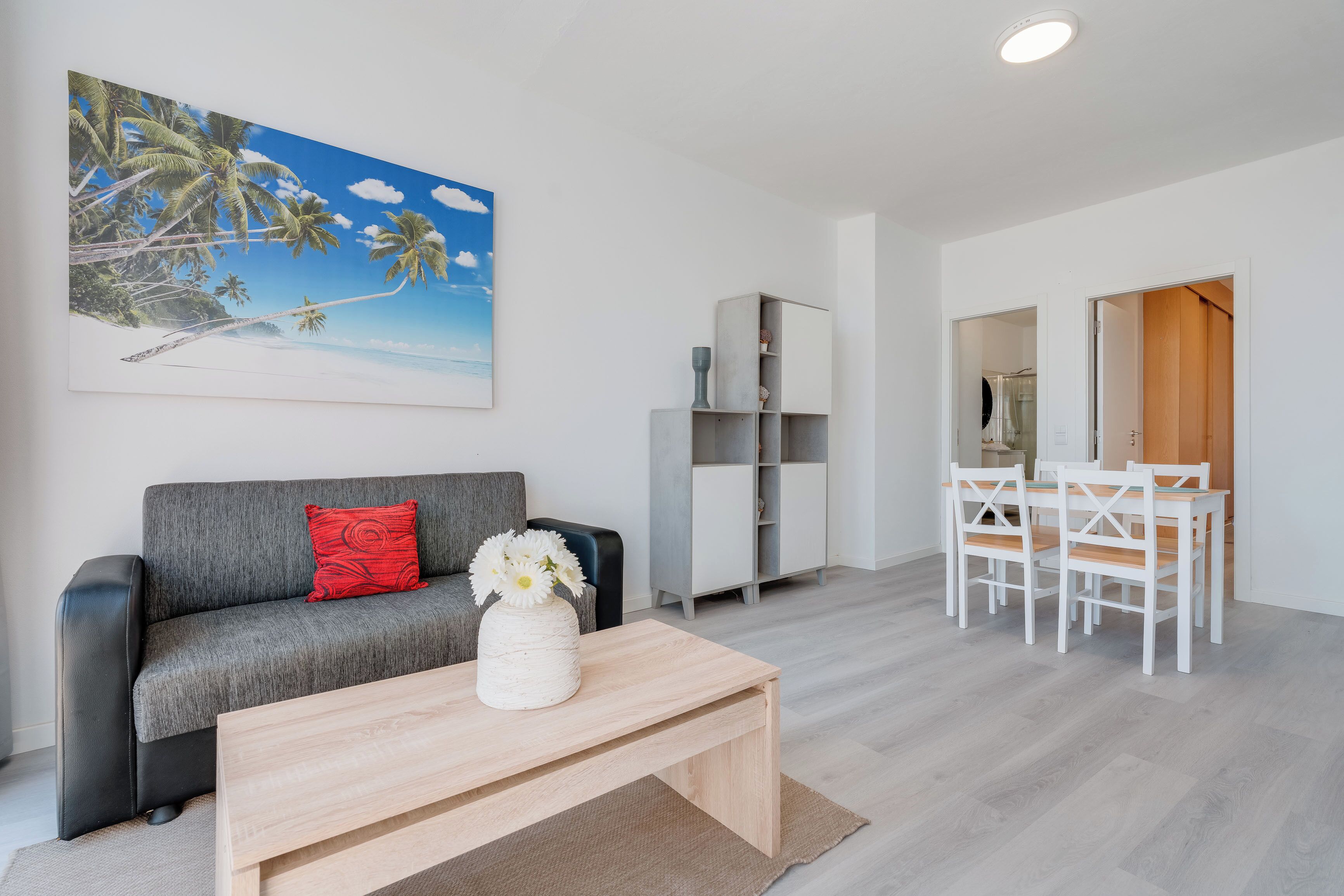 Una sala de estar moderna con un sofá gris, una mesa de centro blanca, un cuadro de una playa, un mueble gris y un comedor con una mesa y sillas blancas.