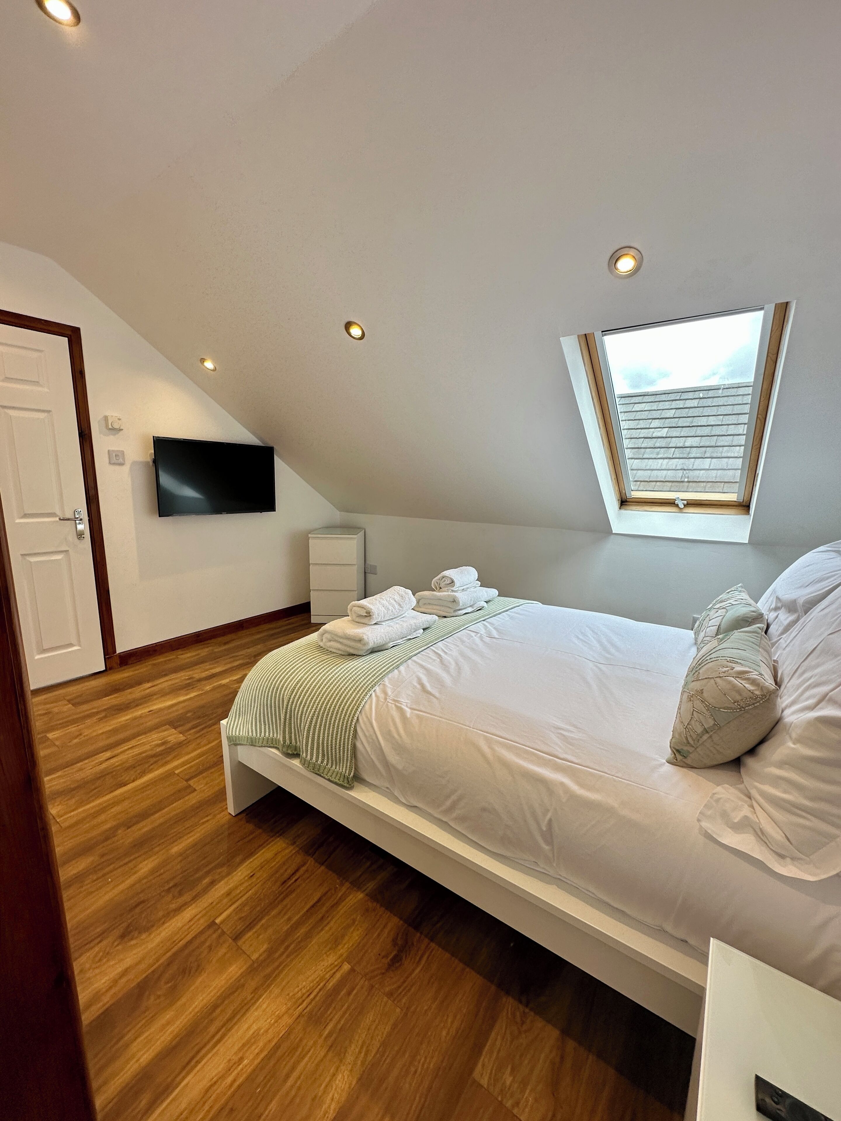A bedroom with a sloped ceiling, a skylight, a bed with white linens, a flat-screen TV, and a wooden floor.