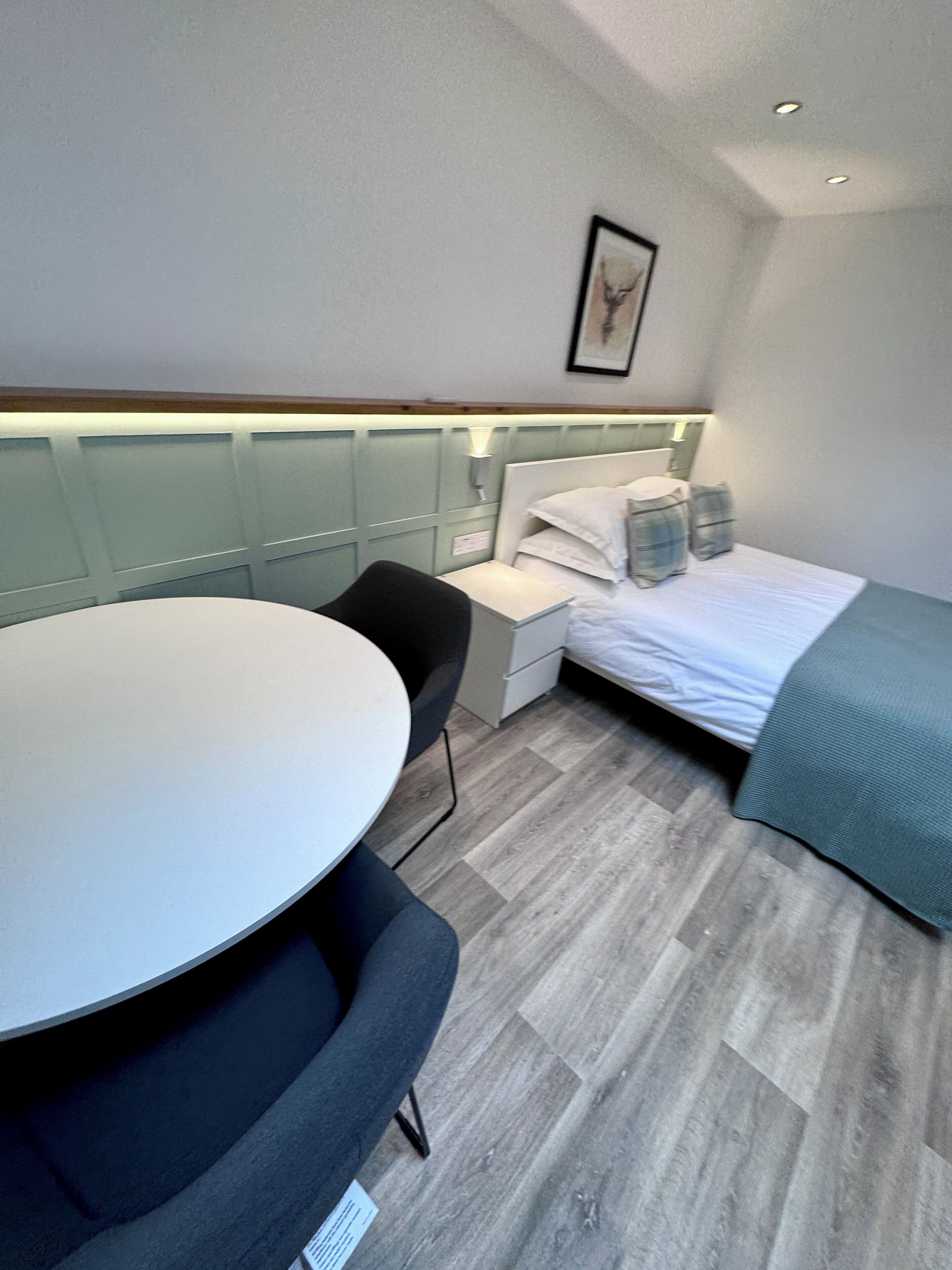 Quarto de hotel pequeno, com cama, mesa de cabeceira, cadeira e uma pequena escrivaninha com cadeira.