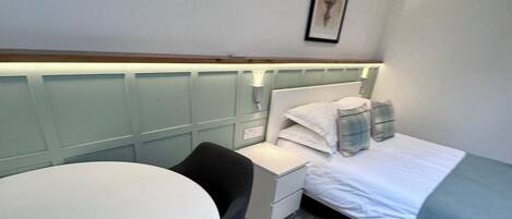 Quarto de hotel pequeno, com cama, mesa de cabeceira, cadeira e uma pequena escrivaninha com cadeira.