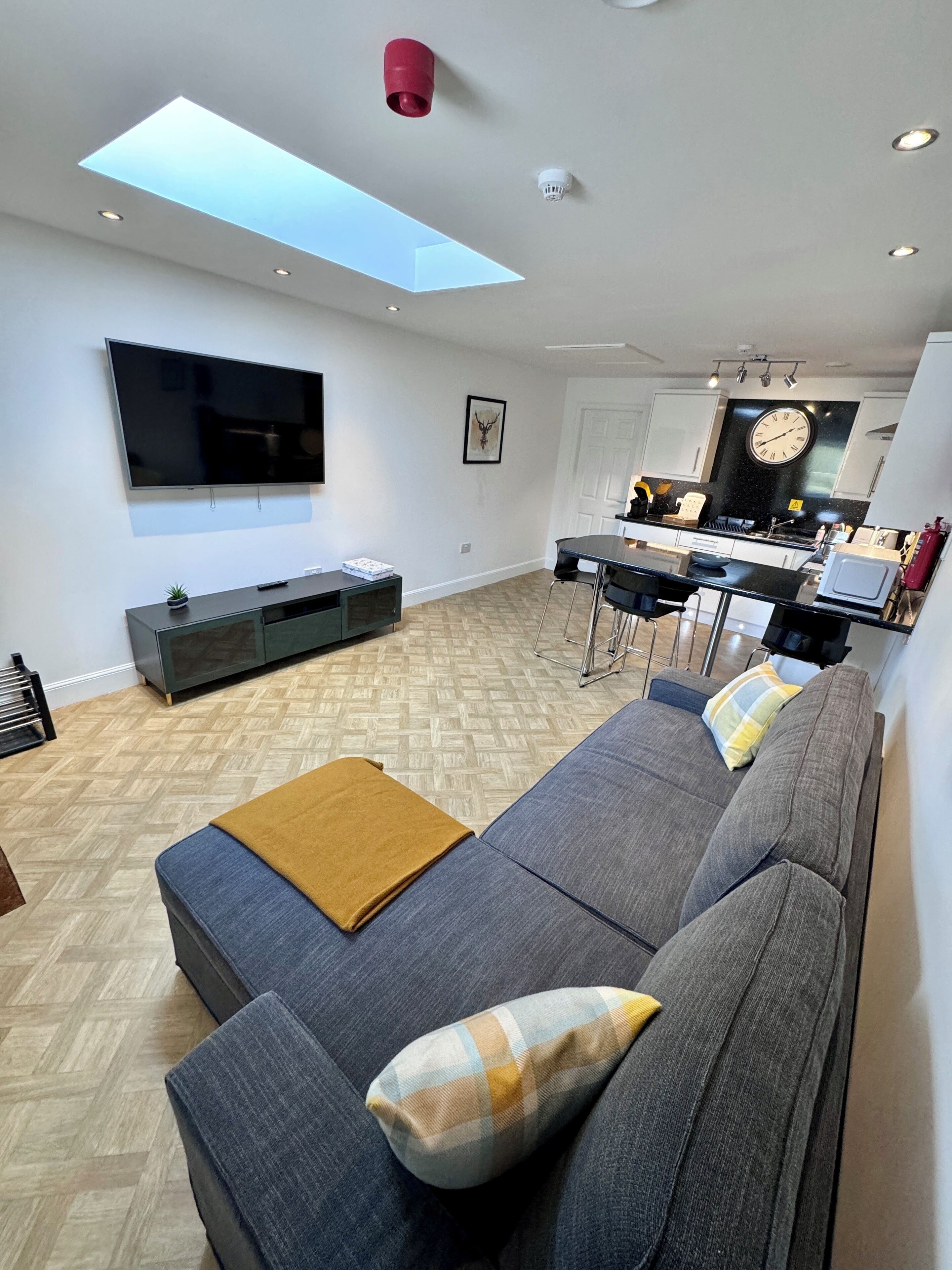 A modern living room with a flat-screen TV, a sofa, a coffee table, and a kitchen area with a clock and a stove.