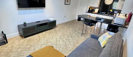 A modern living room with a flat-screen TV, a sofa, a coffee table, and a kitchen area with a clock and a stove.