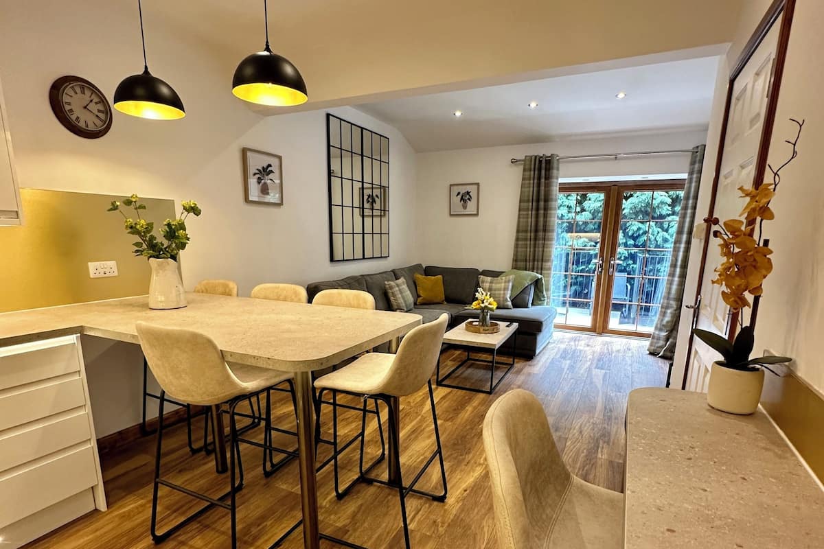 A modern kitchen with a dining area, wooden flooring, pendant lights, and a view through a glass door.