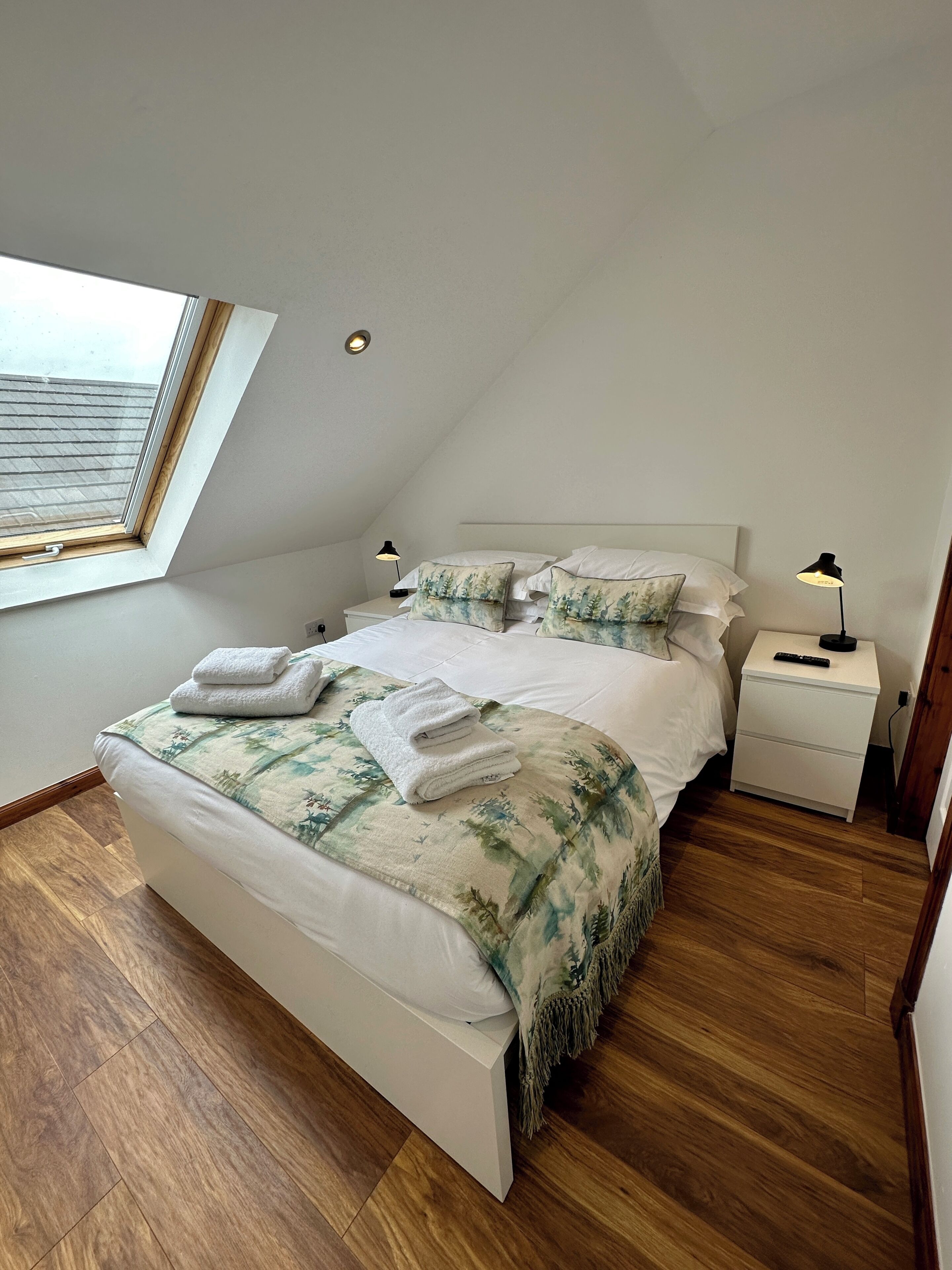 A bedroom with a skylight, a bed with a floral bedspread, two bedside tables with lamps, and wooden flooring.