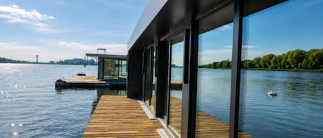 A modern waterfront property with a wooden deck, large glass windows, and a view of a river.