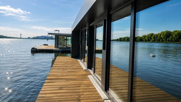Een modern gebouw met grote ramen met uitzicht op een rustig meer, een houten terras en een klein steigertje.