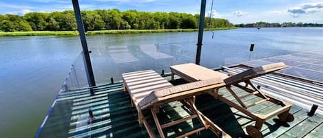 A covered deck with lounge chairs and a table, overlooking a calm body of water.