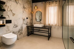 Un baño con inodoro blanco, pared de mármol, tocador negro con espejo y zona de ducha.