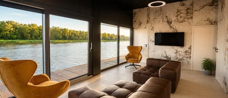 A modern living room with a large window overlooking a body of water, a flat-screen TV mounted on the wall, and a seating area with leather sofas and chairs.
