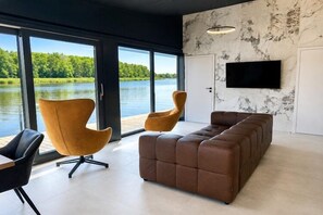 Ein modernes Wohnzimmer mit einer Couch, zwei Sesseln, einem Fernseher und großen Fenstern mit Blick auf einen See.