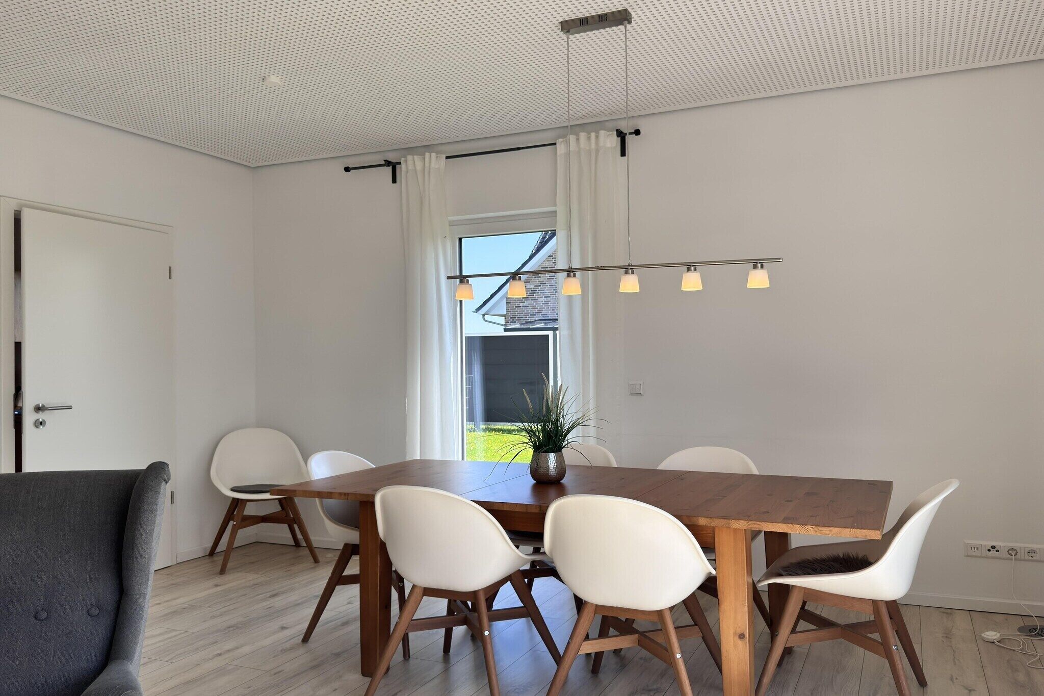 A dining area with a wooden table, white chairs, a grey sofa, and a ceiling light fixture.