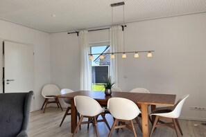 Ein Essbereich mit Holztisch, weißen Stühlen, einem grauen Sofa und einer Deckenleuchte.