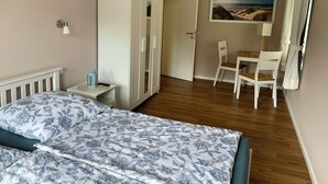 A bedroom with a bed, a wardrobe, a desk with a chair, a painting on the wall, and a nightstand with a lamp.