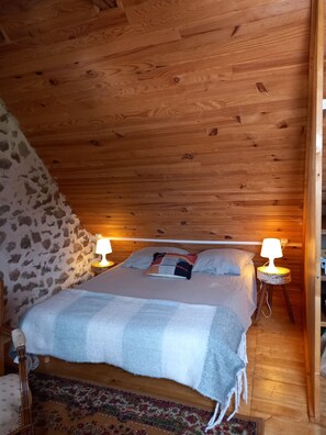 Ein gemütliches Schlafzimmer mit Holzbalkendecke, Steinwand, einem Bett mit kariertem Überwurf und zwei Nachttischlampen.