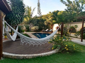 Una hamaca en una terraza de madera, una piscina y abundante vegetación.