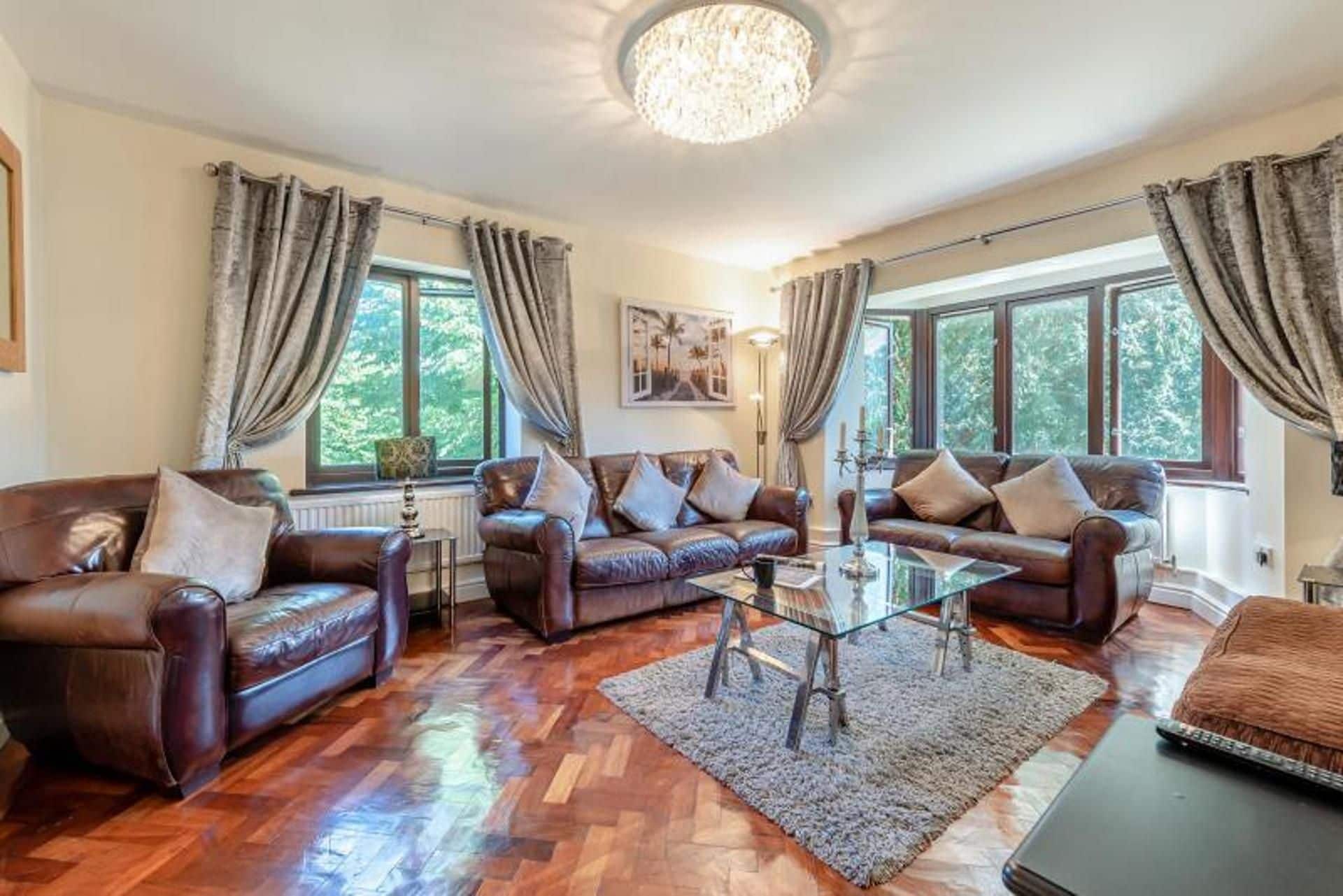 A spacious living room with leather sofas, a glass coffee table, and large windows with curtains.
